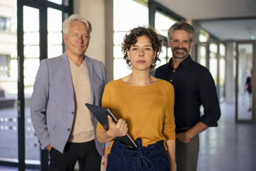 Confident young businesswoman standing with laptop and colleagues in office - JOSEF21756