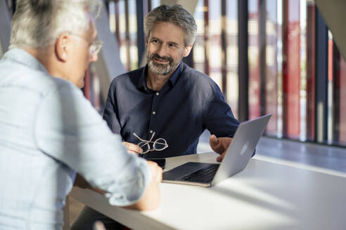 Lächelnder älterer Geschäftsmann, der mit einem Kollegen über einem Laptop am Tisch diskutiert - JOSEF21807