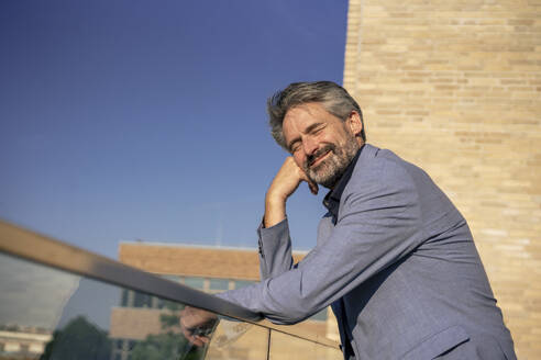 Happy senior businessman with eyes closed leaning on railing - JOSEF21820