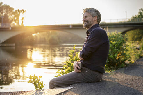 Smiling man sitting with smart phone near lake at sunset - JOSEF21828