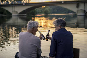 Ältere Freunde stoßen bei Sonnenuntergang mit Bierflaschen am Wasser an - JOSEF21835