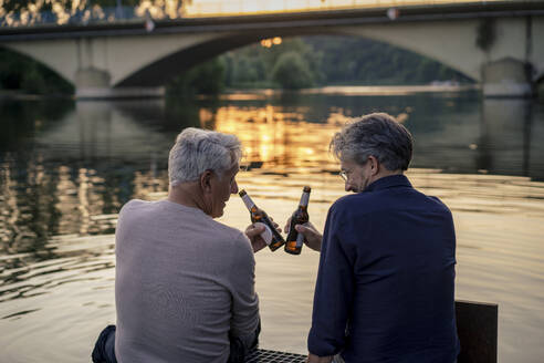 Senior friends toasting beer bottles near water at sunset - JOSEF21835