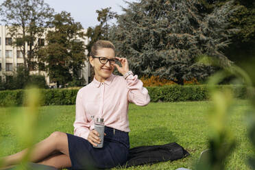 Lächelnde Geschäftsfrau, die ihre Brille im Gras sitzend im Park zurechtrückt - OSF02288