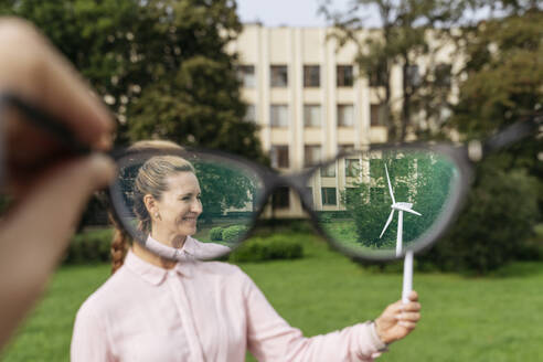 Lächelnde Geschäftsfrau, die ein Modell einer Windkraftanlage hält, gesehen durch eine konkave Brille im Park - OSF02293