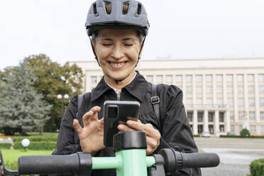 Lächelnde Frau, die mit einem Elektroroller steht und ein Smartphone im Park benutzt - OSF02296