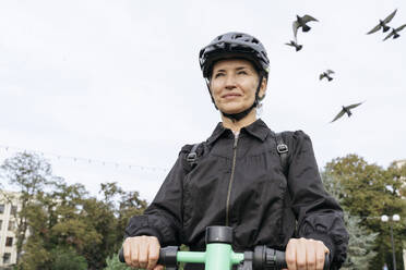 Glückliche Frau trägt einen Helm und fährt einen Elektroroller im Park unter dem Himmel - OSF02297