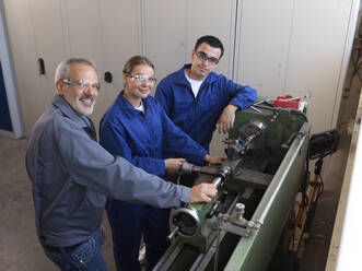 Smiling instructor standing with apprentices near lathe machine at workshop - CVF02615