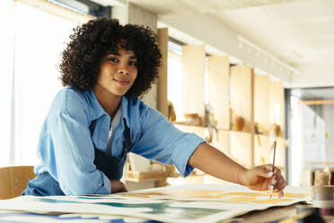 Künstlerin mit lockigem Haar malt mit Pinsel am Schreibtisch im Atelier - EBSF03967