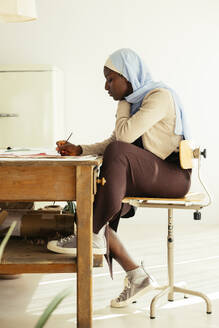 Künstlerin mit Kopftuch malt mit Pinsel am Schreibtisch im Atelier - EBSF03968