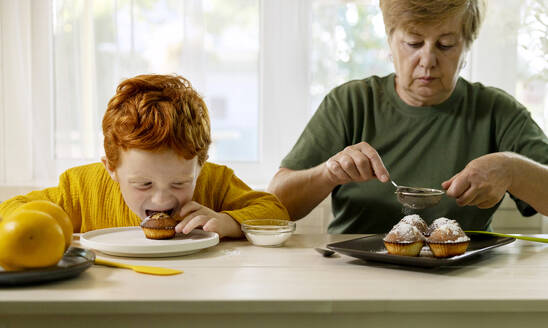 Großmutter bestäubt Muffins mit Zucker und Enkel isst in der Küche zu Hause - MBLF00043