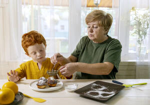 Großmutter streut Zucker auf Muffins für Enkel in der Küche - MBLF00045
