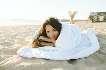 Smiling woman wrapped in blanket lying at beach - SIF01043