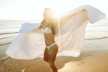 Verspielte Frau im Bikini und mit Decke am Strand drehend - SIF01057