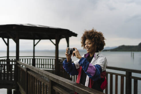 Glückliche junge Frau beim Fotografieren mit Kamera vor einem See - BOYF02019