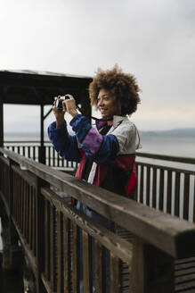 Happy young woman photographing through camera on footbridge - BOYF02020