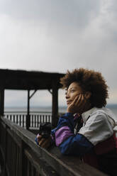 Nachdenkliche junge Frau steht mit Kamera am Geländer - BOYF02022