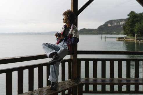 Thoughtful young woman sitting on railing in front of lake - BOYF02033