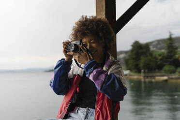 Lächelnde junge Frau beim Fotografieren mit Kamera in der Nähe eines Sees - BOYF02034