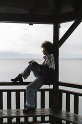 Young woman writing in diary sitting on railing under gazebo - BOYF02039