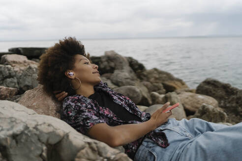 Smiling young woman relaxing on rocks and listening to music through wireless In-ear headphones - BOYF02052
