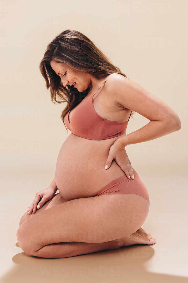 Happy pregnant woman kneeling on the floor in a studio, smiling at her  beautiful baby bump. Young mom-to-be wearing lingerie, cherishing the life  growing inside her with maternal love. stock photo