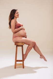 Pregnant woman cradling her belly in a studio. She sits on a chair, dressed in underwear, displaying her prenatal body proudly. Woman radiating positivity and celebrating the journey of motherhood. - JLPSF30811