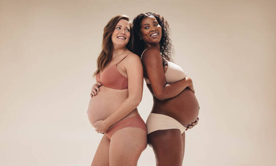 Two confident, pregnant women standing together in a studio. They proudly showcase their baby bumps, smiling and radiating happiness. These moms-to-be celebrate the beauty of their pregnancy bodies. - JLPSF30826