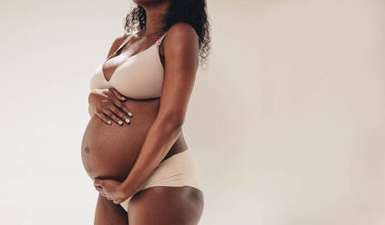 Eine schwangere Afrikanerin im dritten Trimester steht selbstbewusst in einem Studio und umarmt ihren Babybauch. Eine dunkelhäutige schwangere Frau trägt Unterwäsche und umarmt stolz ihre Körperveränderungen mit Liebe. - JLPSF30837