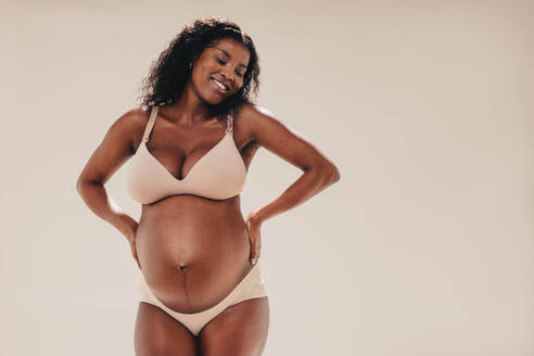 Eine glückliche, selbstbewusste schwarze Frau im dritten Trimester steht in einem Studio und zeigt ihren schönen Babybauch. Sie trägt Unterwäsche und umarmt ihre melaninreiche Haut mit einem Lächeln und Positivität. - JLPSF30838