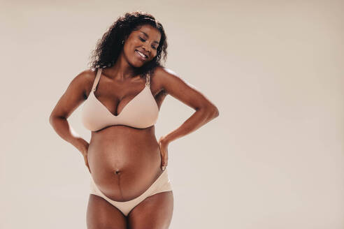 Happy, confident black woman in her third trimester stands in a studio, showing off her beautiful baby bump. She wears lingerie, embracing her melanin-rich skin with a smile and positivity. - JLPSF30838