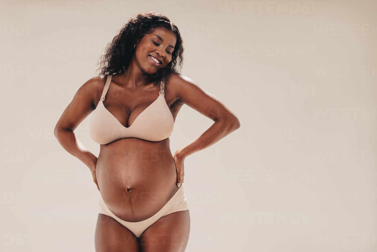 Happy, confident black woman in her third trimester stands in a studio,  showing off her beautiful baby bump. She wears lingerie, embracing her  melanin-rich skin with a smile and positivity. stock photo