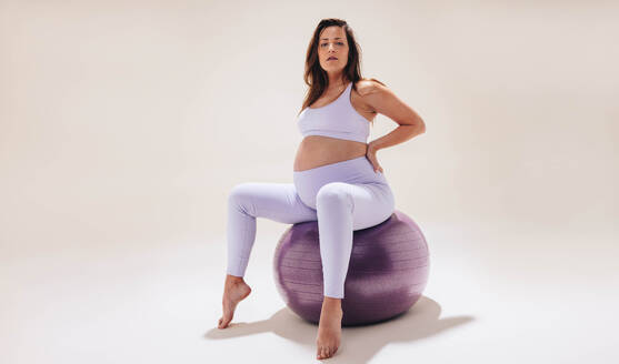 Junge Frau übt in einem Studio Schwangerschaftsyoga auf einem Stabilitätsball. In Fitnesskleidung präsentiert sie ihren Babybauch mit Kraft und Flexibilität. Sie achtet auf ihr Wohlbefinden und ihre Schwangerschaftsvorsorge und blickt gelassen in die Kamera, während sie die Reise der Mutterschaft begrüßt. - JLPSF30851