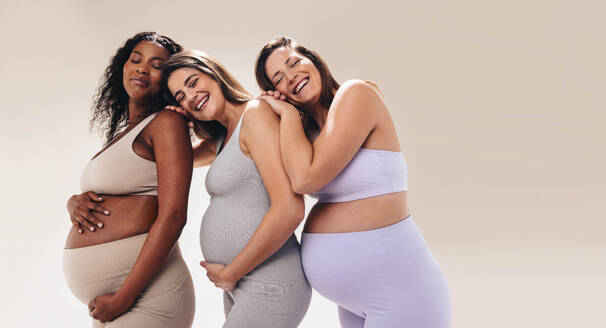 Multikulturelle schwangere Frauen in Fitnesskleidung besuchen eine pränatale Yogastunde und stützen sich gegenseitig. Glücklich und lächelnd legen sie Wert auf Selbstfürsorge und Wohlbefinden während ihrer Schwangerschaft. - JLPSF30869
