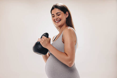 Eine schwangere Frau im dritten Trimester hebt in einem gut ausgestatteten Fitnessstudio eine Kettlebell. Sie legt Wert auf ihr Wohlbefinden und geht mit einer glücklichen und fitten Einstellung an die Mutterschaft heran. - JLPSF30879