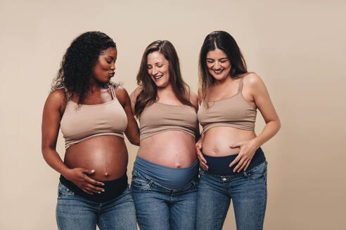 Eine Gruppe von werdenden Müttern zeigt in einem Studio stolz ihren Babybauch. In Jeans und BHs lächeln diese jungen Frauen im dritten Trimester zusammen, verkörpern die Freude der Mutterschaft und feiern die Schönheit der Schwangerschaft. - JLPSF30886