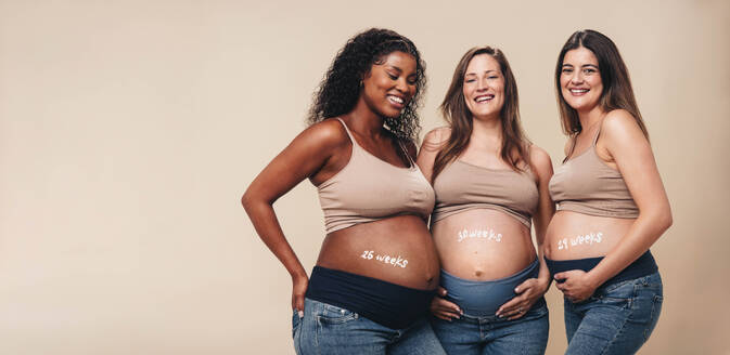 Eine Gruppe schwangerer Frauen im dritten Trimester steht in einem Studio zusammen. Sie tragen Jeans und BHs und zeigen stolz ihren Babybauch. Diese werdenden Mütter aus verschiedenen Kulturen teilen die Freude an der Schwangerschaft. - JLPSF30888