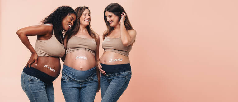 Eine Gruppe von werdenden Müttern im dritten Schwangerschaftsdrittel steht zusammen und unterstützt sich gegenseitig mit einem freudigen Lächeln. Stolz zeigen sie ihren Babybauch und freuen sich mit ihren Freundinnen über die Schwangerschaft. - JLPSF30889