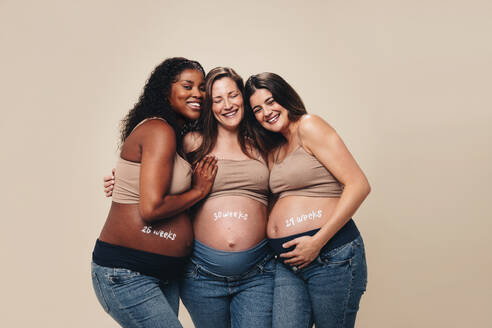 Werdende Mütter stehen in einem Studio zusammen, umarmen sich und unterstützen sich gegenseitig während der Schwangerschaft. Unterschiedliche Frauen lächeln und feiern ihre Babybäuche in bequemen Umstandsjeans und BHs. - JLPSF30891