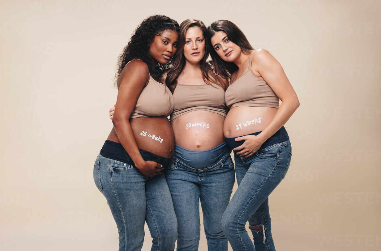 Young, diverse pregnant women in the third trimester stand together in a  studio. They wear jeans and bras, showcasing their baby bumps. Hugging and  looking at the camera, they represent a supportive