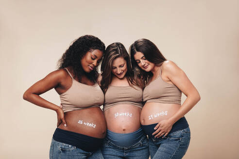 Werdende Mütter im dritten Trimester stehen in einem Studio zusammen und lächeln auf ihre schwangeren Bäuche. Diese jungen Frauen, die Jeans und BHs tragen, unterstützen sich gegenseitig und zeigen die Stärke der Mutterschaft. - JLPSF30895