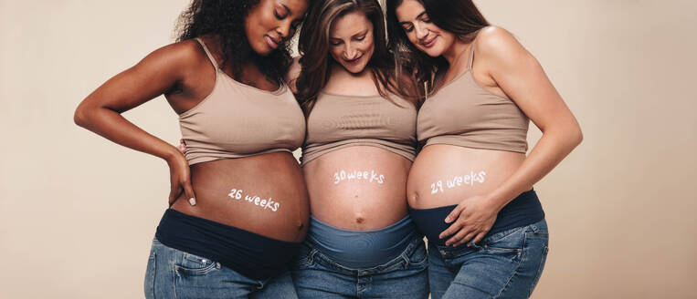 Verschiedene schwangere Frauen im dritten Trimester stehen zusammen und lächeln über ihren Babybauch. Sie unterstützen sich gegenseitig in einem Studio und feiern die Mutterschaft mit Jeans und BHs. Diese jungen Mütter strahlen Wohlbefinden aus und umarmen die Schönheit der Schwangerschaft. - JLPSF30896