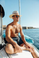 Fit shirtless man in swimming shorts and hat sitting on sailboat enjoying summer vacation against sea and cloudless blue sky - ADSF48508