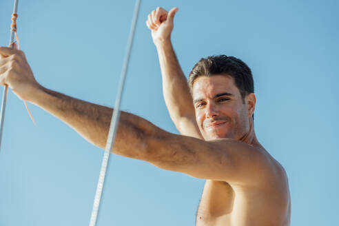 Side view of happy shirtless man in swimming shorts standing on sailboat enjoying summer vacation against cloudless blue sky - ADSF48509