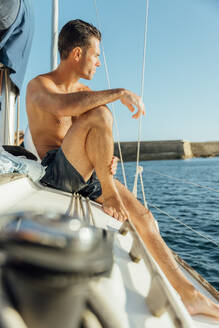 Side view of fit shirtless man in swimming shorts sitting on sailboat enjoying summer vacation against sea and cloudless blue sky - ADSF48510