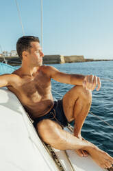 Side view of fit shirtless man in swimming shorts sitting on sailboat enjoying summer vacation against sea and cloudless blue sky - ADSF48511