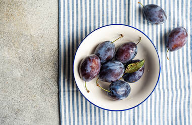 Top view of ripe plums served in ceramic bowl on striped napkin against gray surface - ADSF48515