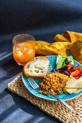 High angle of five ingredients lunch set consists of homemade goat cheese, fresh cucumber and tomato with leaves of basil herb, in addition to fried rice with meat with slices of healthy sour dough bread served with glass of carrot-apple juice against gray background - ADSF48517