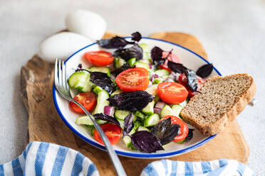 Closeup of Seasonal vegetable salad with organic cucumber, tomato, onion and red basil leaves served on plate placed on wooden tray near napkin against gray background - ADSF48518