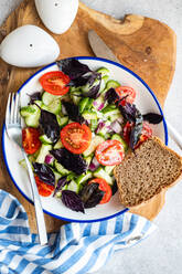 Top view of Seasonal vegetable salad with organic cucumber, tomato, onion and red basil leaves served on plate placed on wooden tray near napkin against gray background - ADSF48519