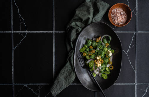 Top view of cucamelon salad with red onion, olives and spicy cheese served on black plate with fork near napkin on dark table with white lines - ADSF48525